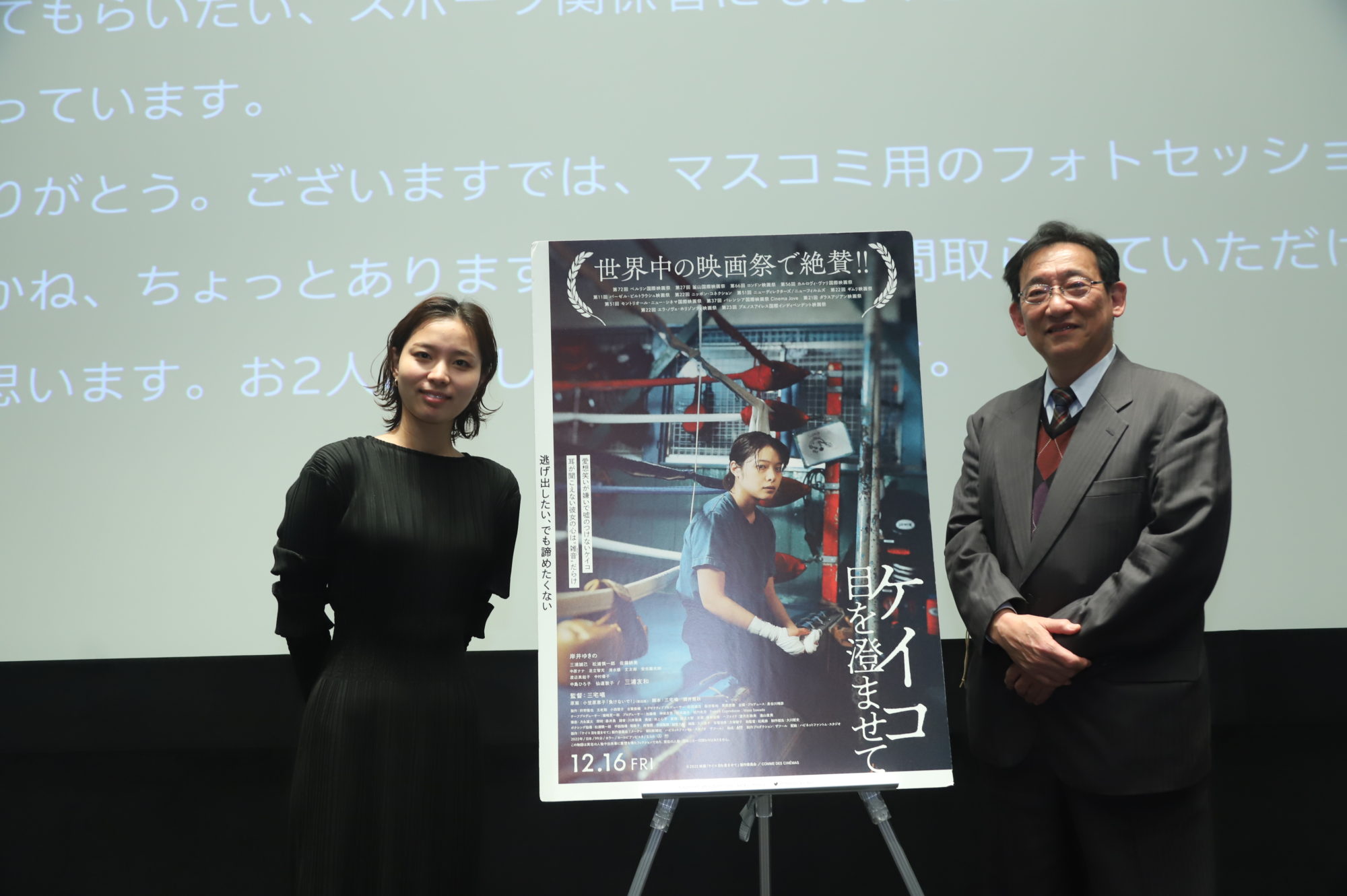 長井さんと越智さん　ケイコ目を澄ませてのポスターを挟んで壇上に立っている
