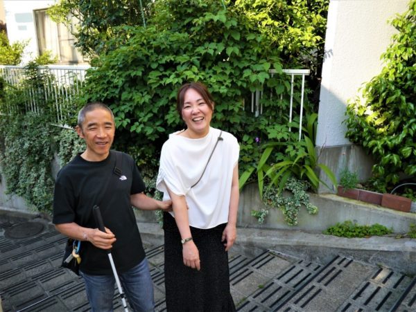 画像:建物裏の階段にて　白杖を持つ小林さんと松田　膝まで映った広めの2ショット　2人とも笑っている