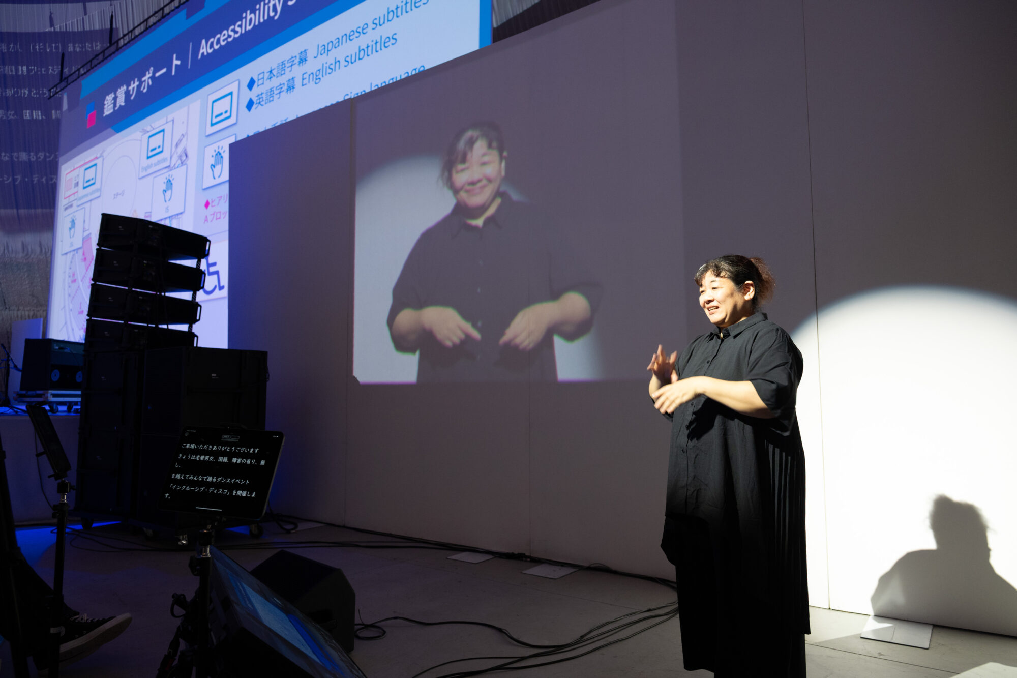 右に手話通訳の方が立っており、手話がスクリーンに映っている。左端にはスクリーンがあり、インフォメーションや字幕が表示されている。